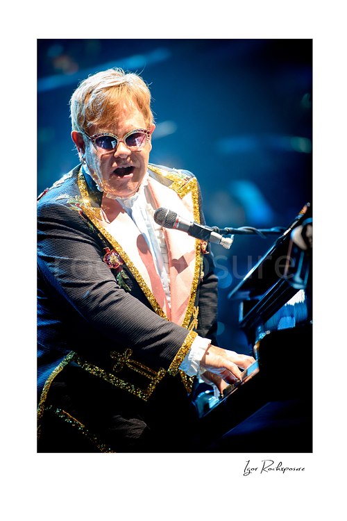 Vertical color image of Elton John singing at the piano in a sequined stage jacket and tinted glasses