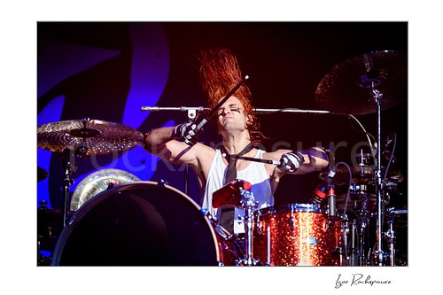 Horizontal color image of Arejay Hale of Halestorm playing drums in a dramatic live performance shot, with drumsticks crossed, red hair flying upward, and a sparkling red drum kit under vivid stage light
