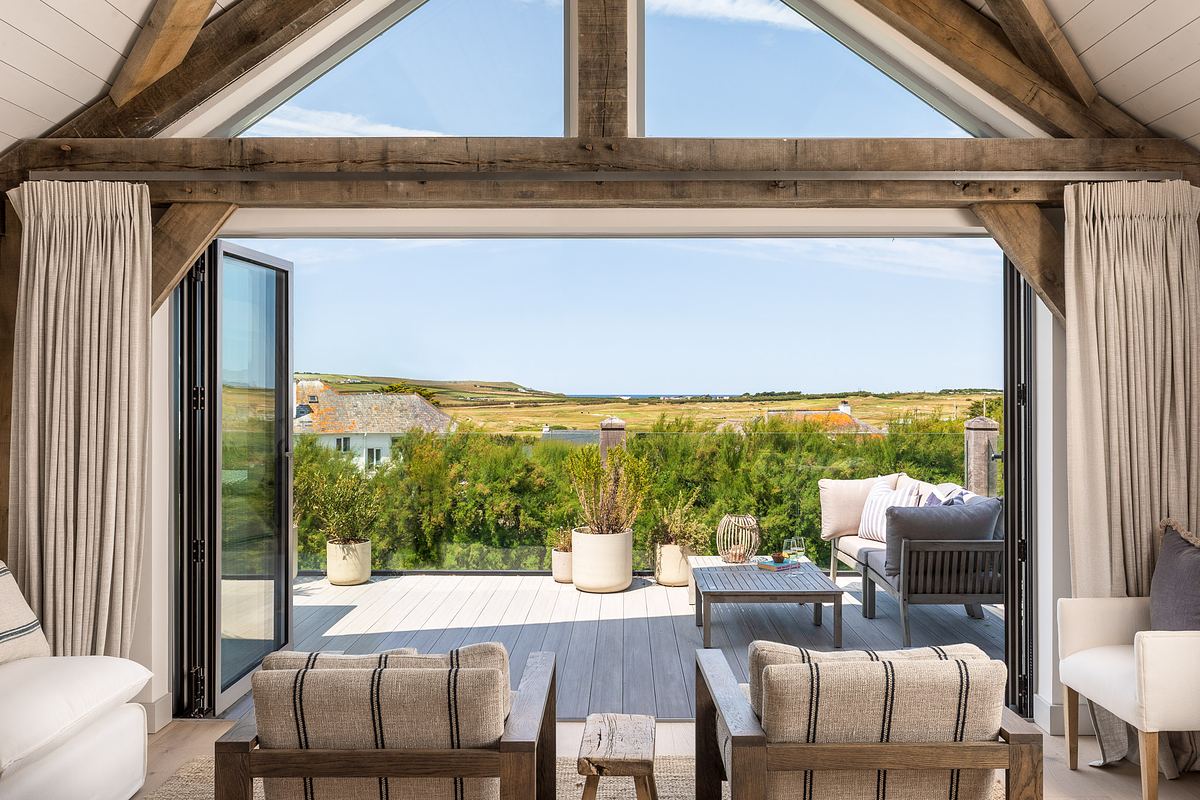 A bright living room with exposed beams opens to a deck with outdoor seating, overlooking grassland and a distant view of the sea under a clear sky.