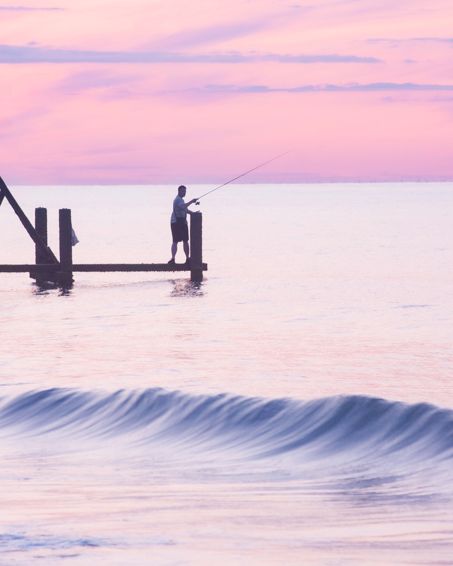 lone fisherman standing on a wooden sea groyne at sunrise on Happisburgh beach with soft pink waves rolling in
