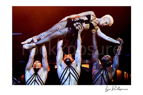 Horizontal color image of Pink held aloft by three dancers during a dramatic live performance under warm red concert light