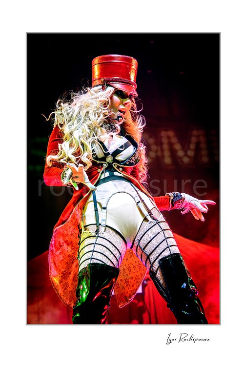 Vertical color image of Maria Brink of In This Moment in a dramatic live performance pose, wearing a red military-style outfit and hat with flowing blonde hair, stage smoke, and vivid red light behind her