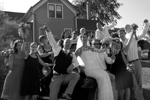 The bridal party lifts their arms in celebration as they sit with the bride and groom on a vintage car