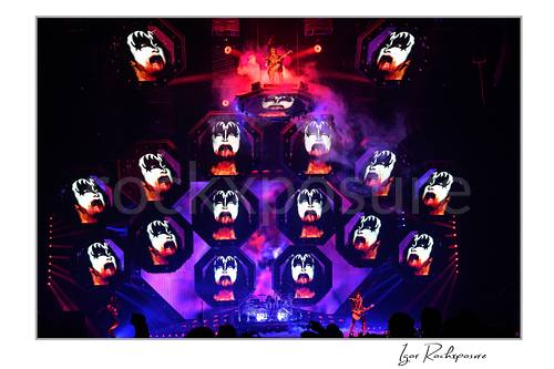 Gene Simmons of KISS standing on an elevated stage platform surrounded by large illuminated Gene Simmons demon face visuals across the stage screens under red and purple concert lighting