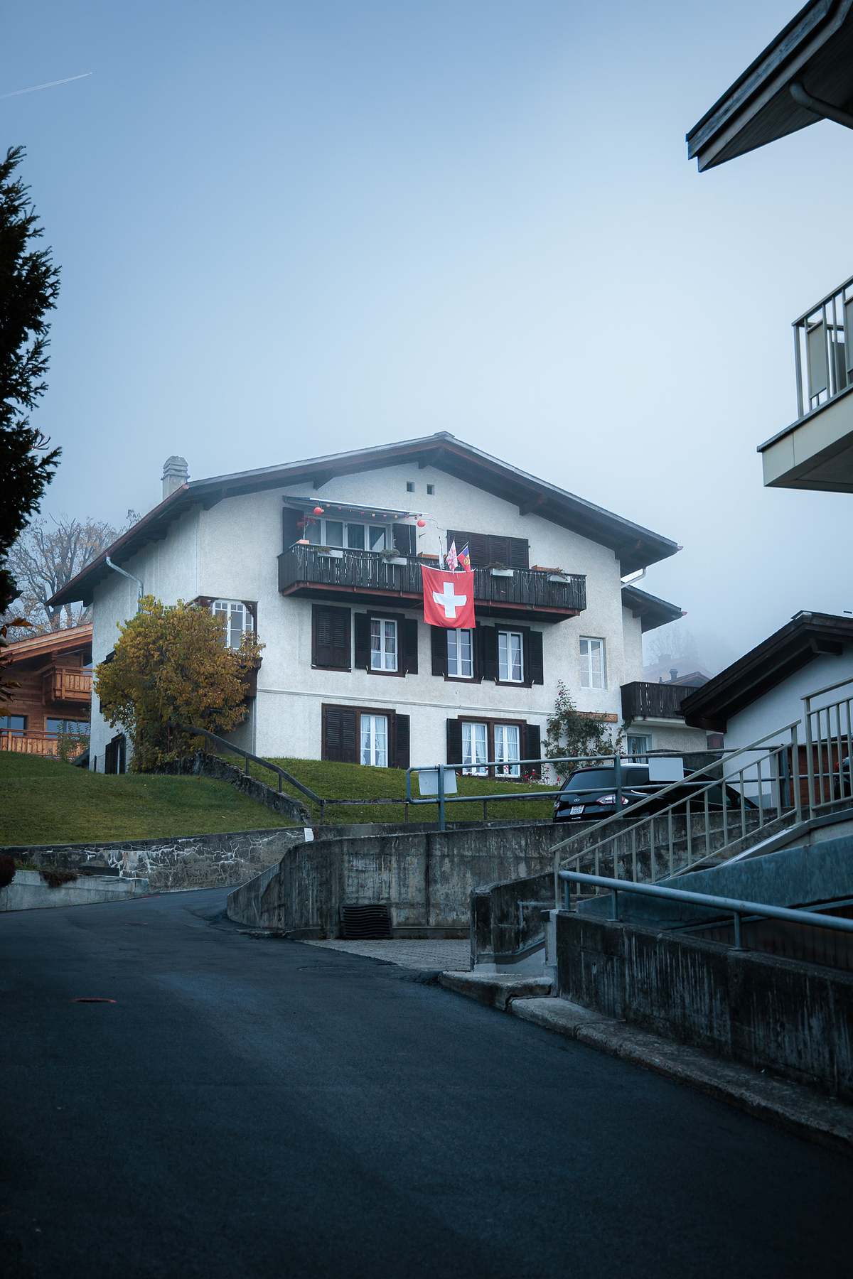 Weisses Schweizer Haus mit grosser Schweizerflagge am Balkon, umgeben von herbstlich gef&auml;rbten B&auml;umen und dichtem Nebel in Grindelwald