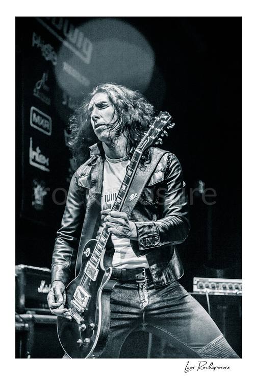 Vertical black and white image of Brent Woods playing guitar on stage wearing a leather jacket in a dramatic performance shot