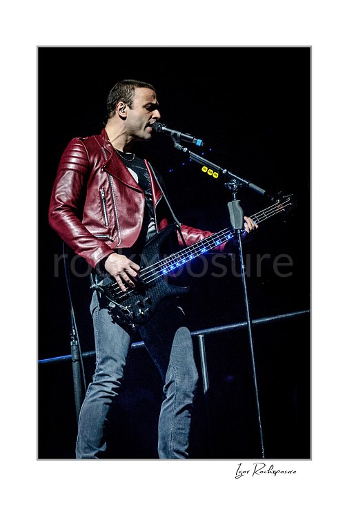 Vertical color image of Chris Wolstenholme of Muse singing into a microphone while playing bass in a red leather jacket