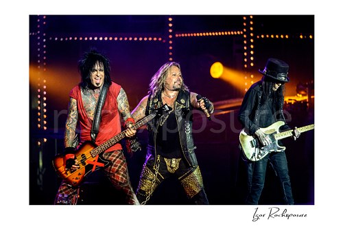 Horizontal color image of Nikki Sixx, Vince Neil and Mick Mars performing live on stage during a Mötley Crüe concert with bass guitar, microphone and warm concert lighting