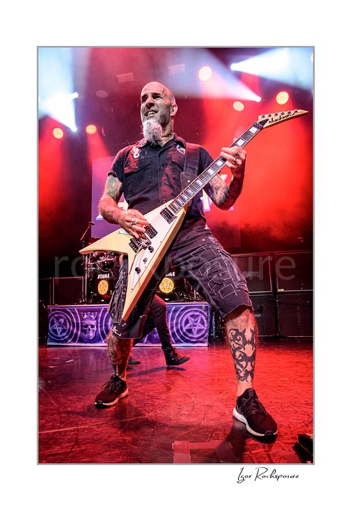 Vertical color image of Scott Ian of Anthrax performing live on stage, playing guitar in a wide stance under vivid red concert lighting