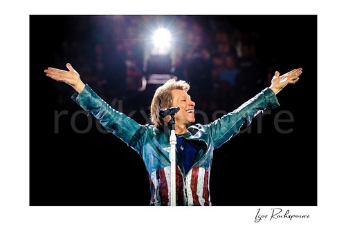 Horizontal color image of Jon Bon Jovi on stage beside a microphone stand with both arms outstretched, smiling under a bright backlight