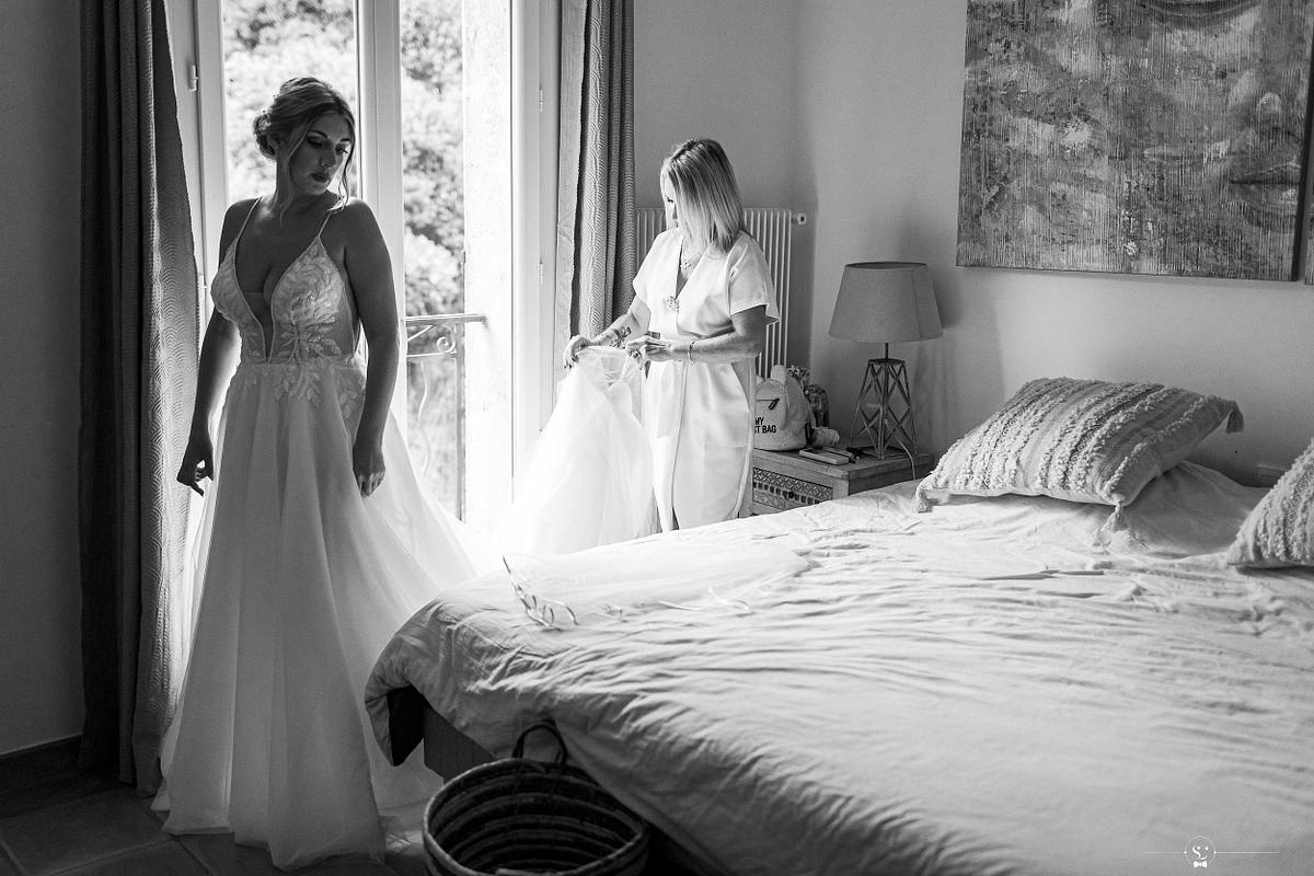 La mari&eacute;e, dans une robe de dentelle fluide, se tient &eacute;l&eacute;gamment &agrave; c&ocirc;t&eacute; d'une porte-fen&ecirc;tre, avec une dame arrangeant sa traine, refl&eacute;tant l'&eacute;l&eacute;gance et la simplicit&eacute; du mariage la&iuml;que &agrave; N&icirc;mes