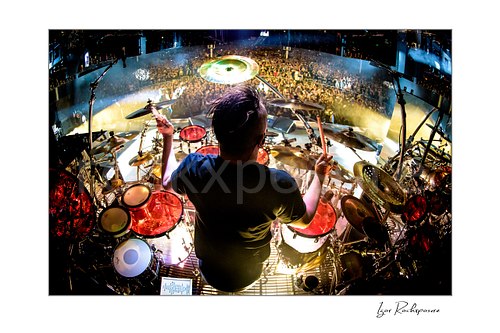 Horizontal color image of Korn drummer Ray Luzier seen from behind at the drums with drumsticks raised above the stage and crowd