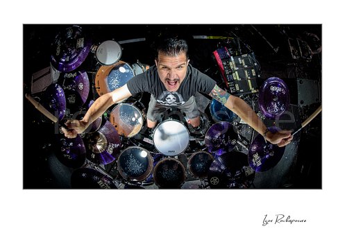 Horizontal color aerial image of Charlie Benante of Anthrax performing live on stage behind his drum kit, photographed from above with drumsticks raised and the full set visible below