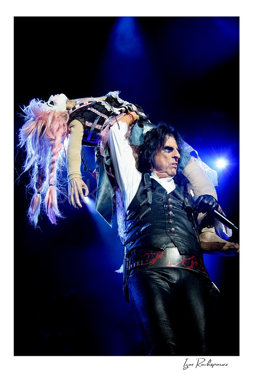 Vertical color image of Alice Cooper performing live on stage while carrying a lifelike female stage prop over his shoulder under blue concert lighting