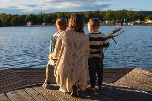 Eine Mutter sitzt von hinten im Gegenlicht mit ihren beiden Söhnen auf einem Holzsteg und blickt über einen weiten See.