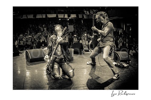 Horizontal black and white image of Tom Keifer and Tony Higbee performing live on stage, with Keifer kneeling in the foreground while Higbee plays beside him and the crowd rises behind them