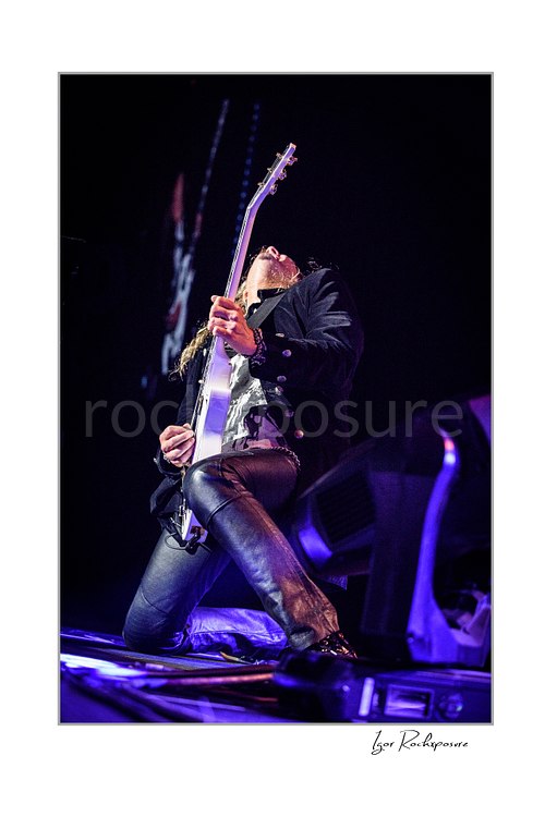 Vertical color low-angle image of Joel Hoekstra of Whitesnake performing live on stage, kneeling while playing electric guitar under purple concert lighting