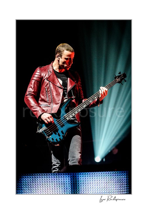 Vertical color image of Chris Wolstenholme of Muse playing bass in a red leather jacket with radiant blue stage beams behind him