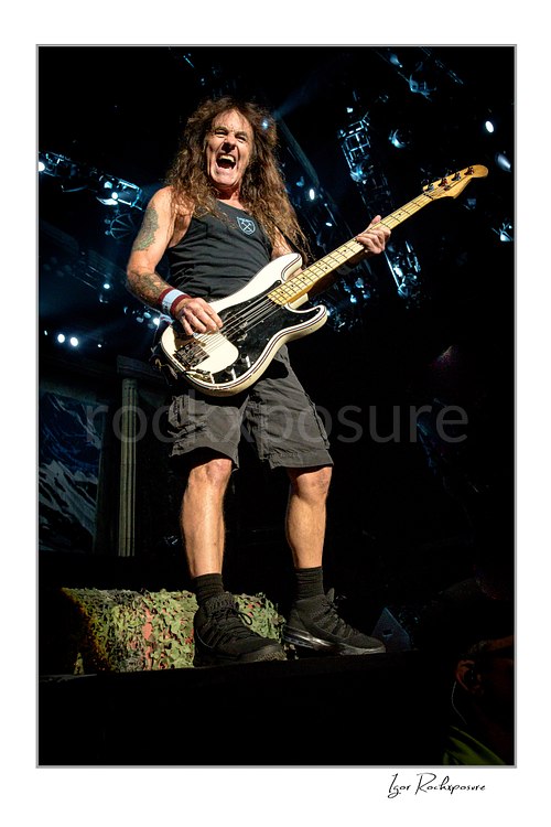 Vertical color image of Steve Harris of Iron Maiden performing live on stage, playing bass guitar under bright concert lighting