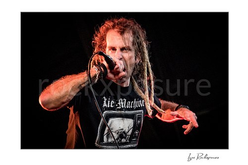 Horizontal color image of Randy Blythe of Lamb of God in a tight stage close-up singing into a handheld microphone with long dreadlocks