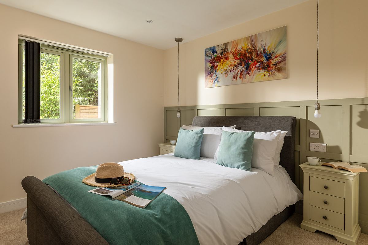 A neatly made bed with teal accents sits between two nightstands in a bright bedroom, styled by a holiday home photographer Cornwall, with a colorful abstract painting above the headboard and light streaming in through a window.