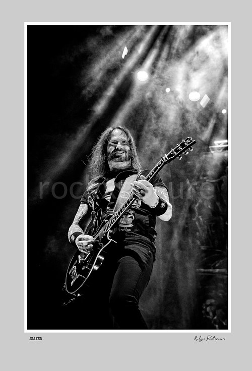 Vertical black and white image of an unidentified guitarist of Slayer playing onstage beneath dramatic overhead concert beams and smoke