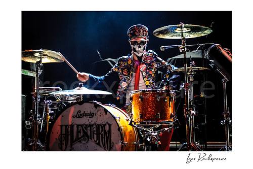 Horizontal color image of Arejay Hale of Halestorm playing drums in a dramatic live performance shot, wearing a skull face covering and a jacket covered in buttons behind a sparkling orange drum kit