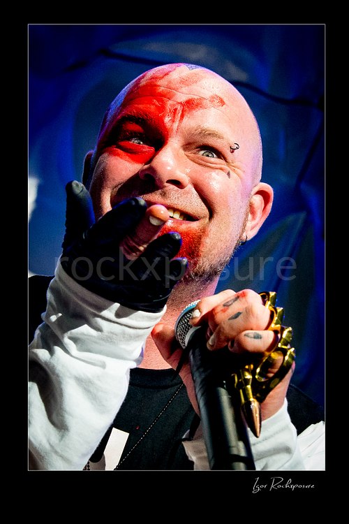 Vertical color image of Ivan Moody of Five Finger Death Punch in a tight onstage close-up holding a microphone under red and blue concert light