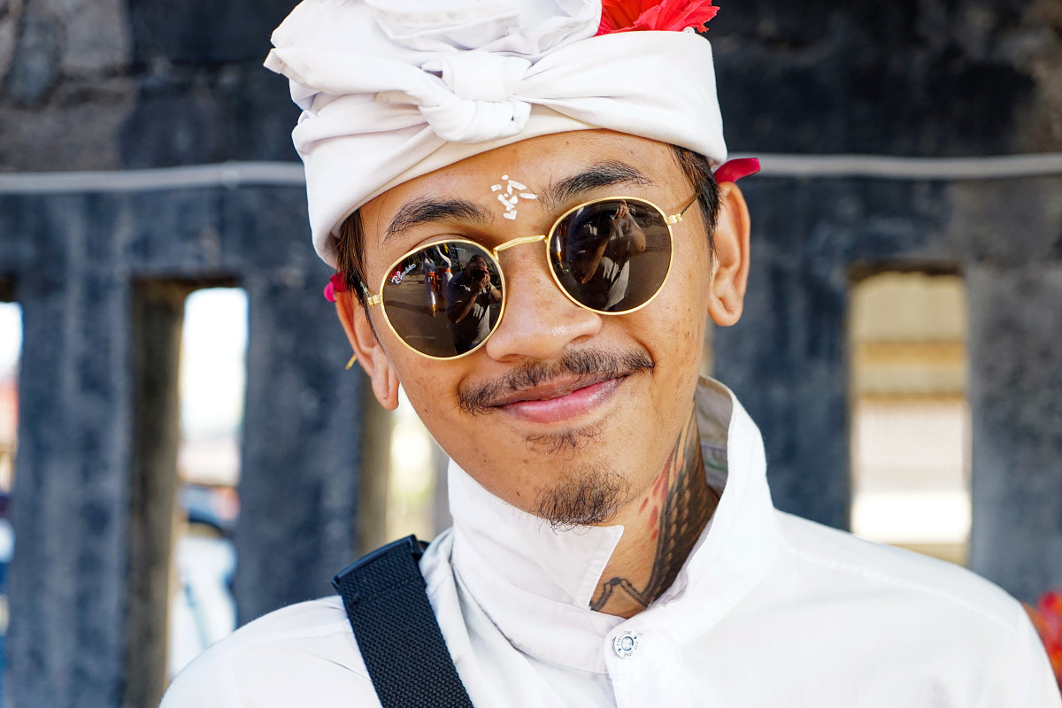 Balinese man wearing traditional ceremonial dress and sunglasses smiling during a religious ceremony in Bali, Indonesia.