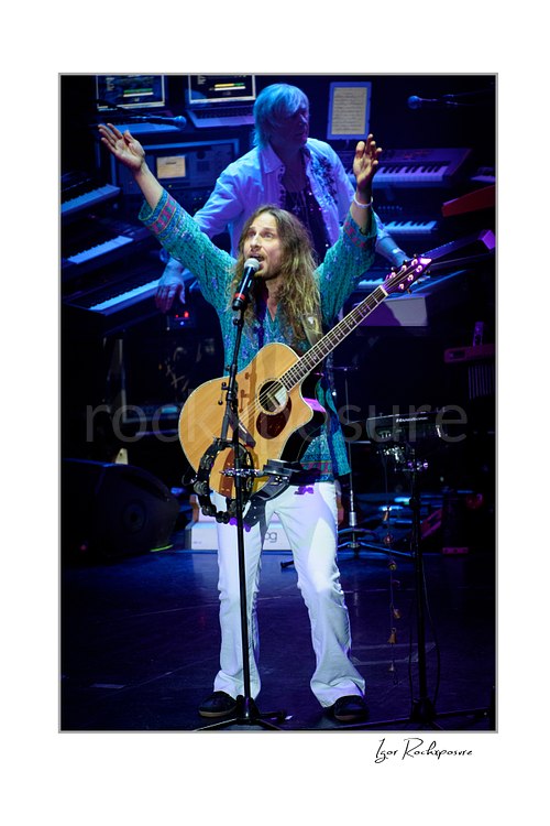 Vertical color image of Jon Davison of Yes singing into a microphone with acoustic guitar and raised arms while Geoff Downes is visible behind him at the keyboard rig