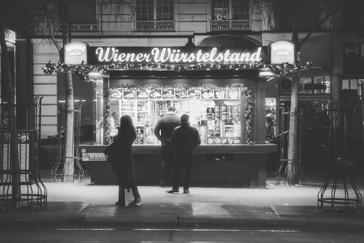 Schwarz-Weiß-Fotografie eines Wiener Würstelstands am Kärntner Ring bei Nacht mit beleuchtetem Verkaufsstand und Menschen davor