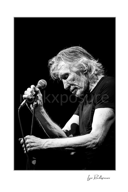 Black and white portrait of Roger Waters looking down, gripping a microphone with one hand and its stand with the other against a black background.