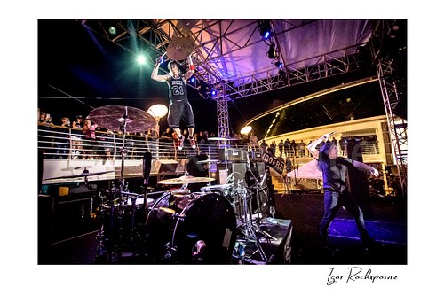 Horizontal color image of Zoltan Chaney leaping above the drum kit while holding a cymbal on stage during a live performance