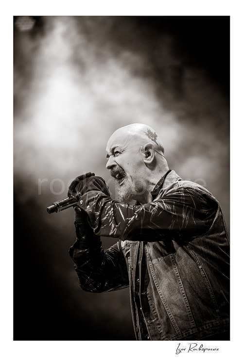 Vertical black and white image of Rob Halford of Judas Priest performing live on stage, singing into a microphone in profile against a smoky background
