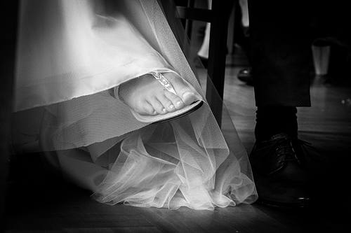 Nahaufnahme unter dem Tisch im Standesamt: Braut hat die Beine überschlagen, ihr eleganter Hochzeits-Flip-Flop und die lackierten Zehen sind durch den Tüll des Kleides sichtbar.