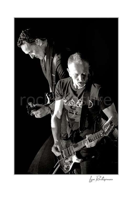 Vertical black and white image of Vivian Campbell and Phil Collen of Def Leppard performing live on stage, with Campbell at left and Collen at right playing guitar against a dark background