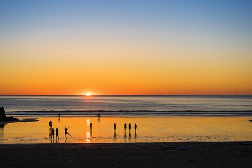 The sun sinks below the horizon at Lusty Glaze beach in Cornwall as people walk towards the shore and a girl dances in silhouette.