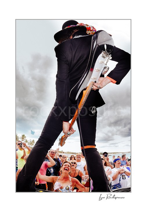 Vertical color image of Randy Hansen standing above a cheering crowd with a guitar in a dramatic low-angle outdoor performance shot
