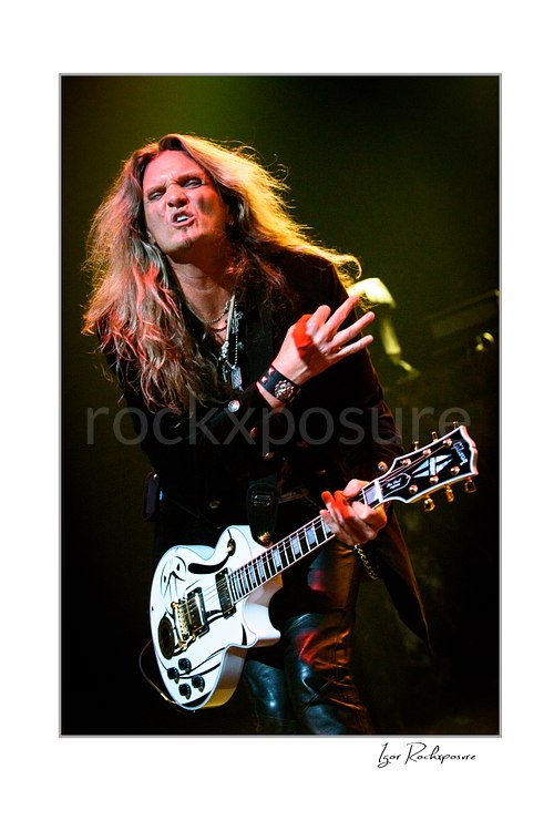 Vertical color image of Joel Hoekstra of Whitesnake performing live on stage, playing electric guitar and gesturing with one hand under warm concert lighting