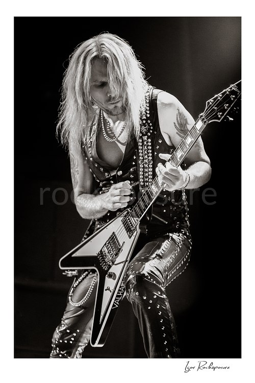 Vertical black and white image of Richie Faulkner of Judas Priest performing live on stage, playing flying V guitar under dramatic concert lighting