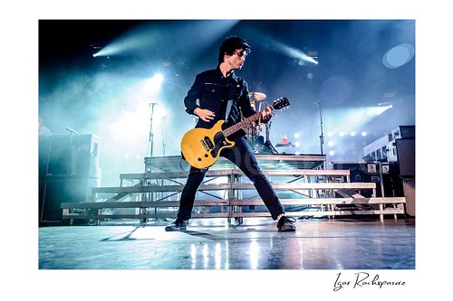 Horizontal color image of Billie Joe Armstrong of Green Day playing guitar onstage in a wide stance under bright concert lights and stage haze