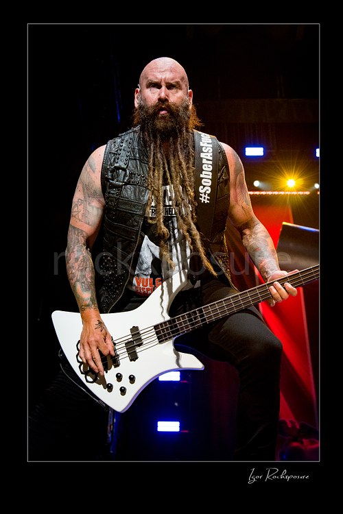 Vertical color image of Chris Kael of Five Finger Death Punch playing bass onstage in a studded vest with long beard and white bass guitar