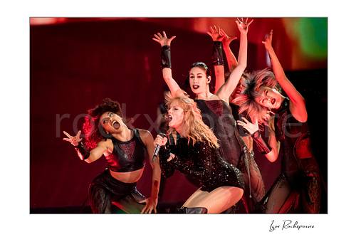 Horizontal color image of Taylor Swift singing on stage with dancers under vivid red concert lighting and dramatic motion