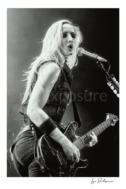 Vertical black and white image of Nita Strauss performing live on stage with an electric guitar beside a microphone