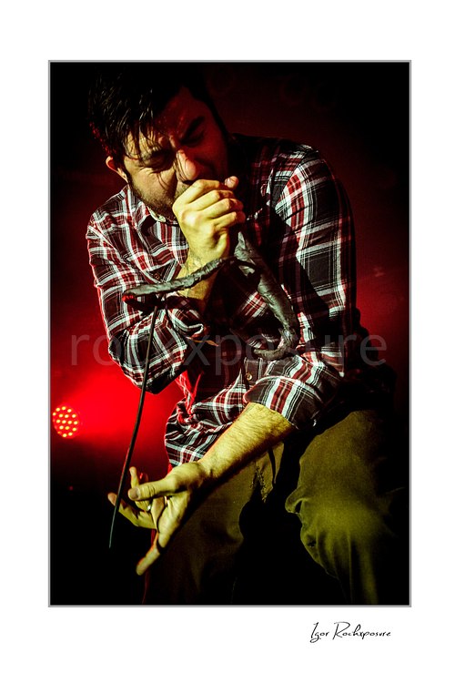 Vertical color image of Chino Moreno of Deftones performing live on stage with a handheld microphone, plaid shirt, wrapped microphone cable, and deep red lighting