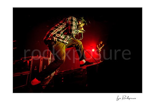 Horizontal color image of Chino Moreno of Deftones performing live on stage in a plaid shirt with a crouched stance, handheld microphone, and intense red lighting