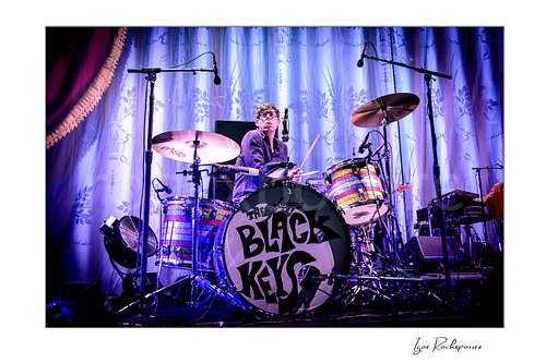 Horizontal color image of Patrick Carney of The Black Keys at the drum kit before a patterned stage curtain under cool purple lighting