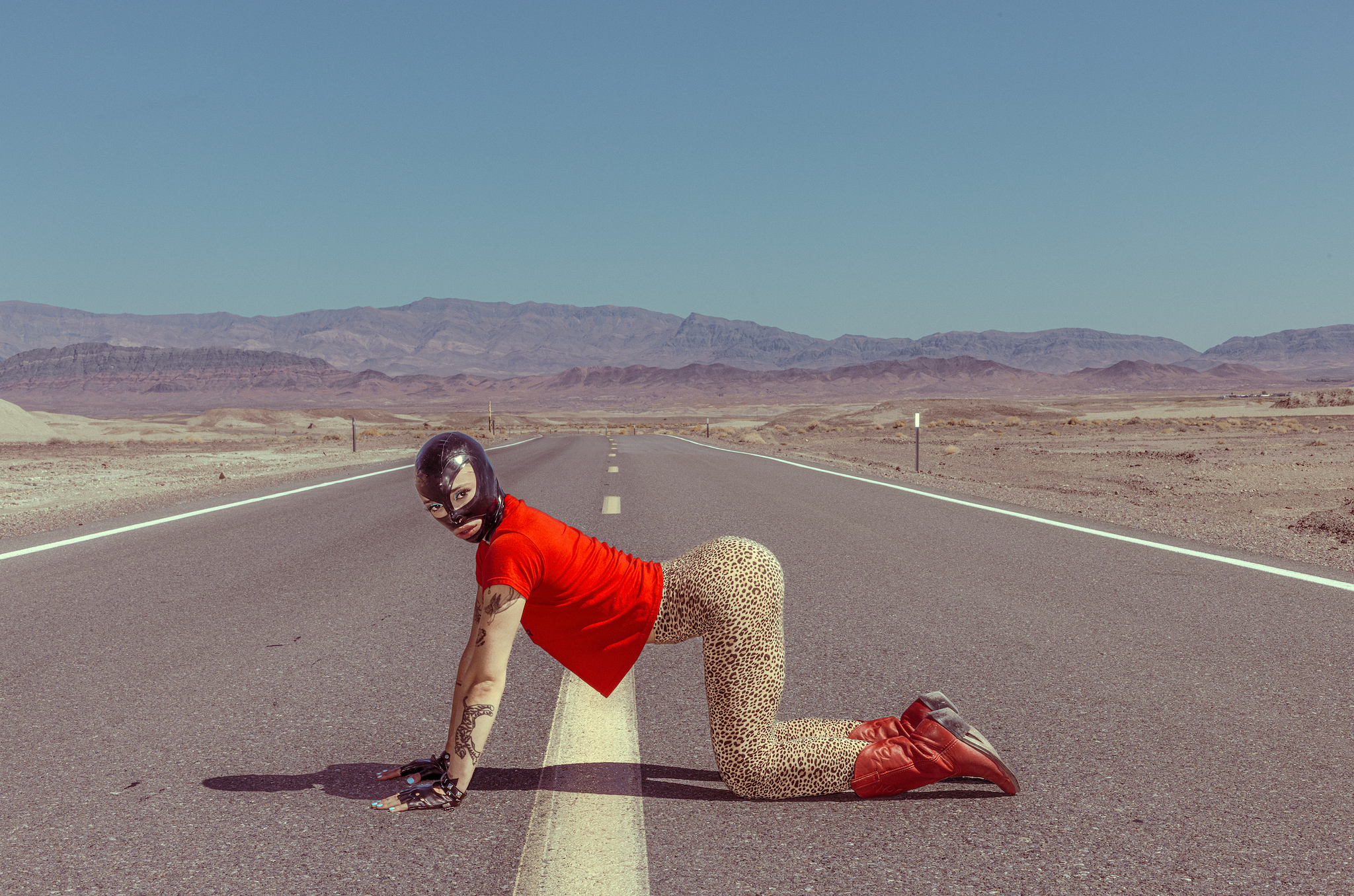 A model on hands and knees in middle of long desert highway in latex mask by copenhagen portrait photographer
