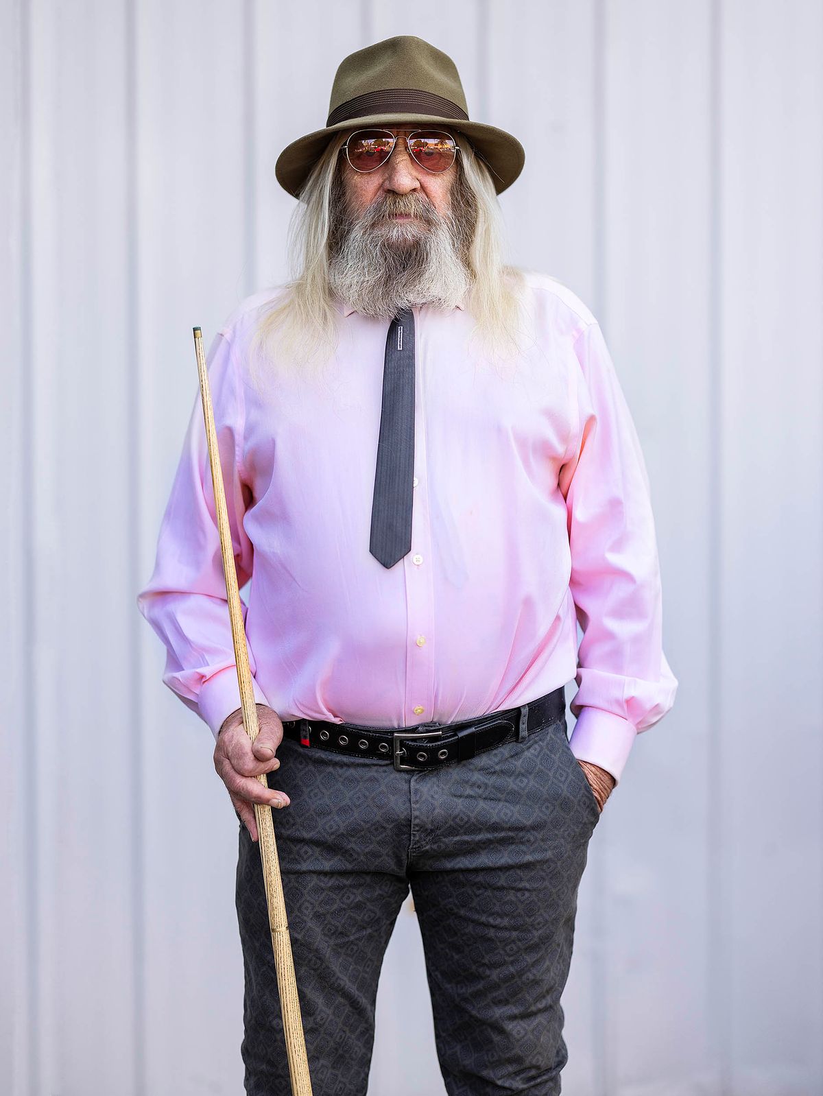 Portrait of a man in pink shirt holding a pool cue, wearing a hat and sunglasses.