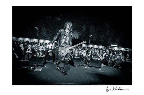 Horizontal black and white image of Tom Keifer performing live in a remote camera shot from the drum kit facing forward, walking across the stage with guitar in hand and the crowd filling the background